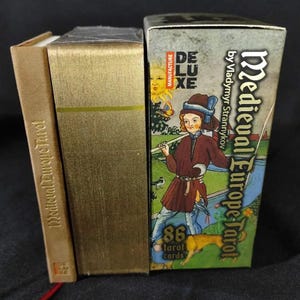 May include: A Medieval Europe Tarot card set with a gold-colored box and a smaller book. The tarot box features a medieval illustration of a person with a dog, the text "Medieval Europe Tarot", and "86 tarot cards".