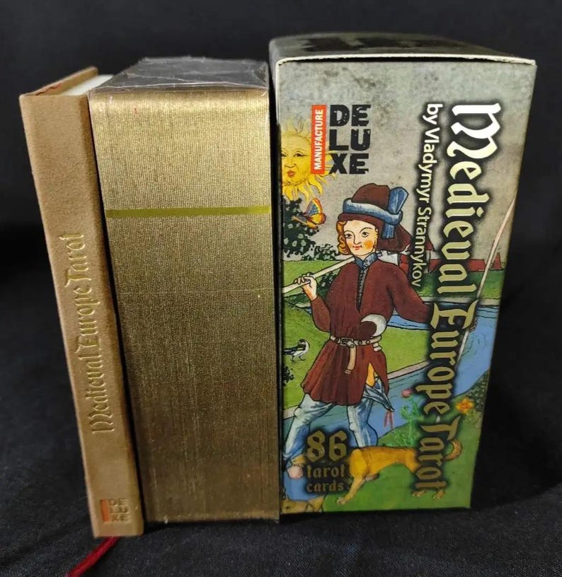 May include: A Medieval Europe Tarot card set with a gold-colored box and a smaller book. The tarot box features a medieval illustration of a person with a dog, the text "Medieval Europe Tarot", and "86 tarot cards".