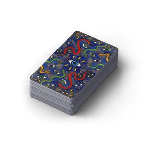 May include: A deck of tarot cards with a blue background and a repeating pattern of eyes and snakes. The center of the deck features a large eye with rays of light. The edges of the cards are silver.