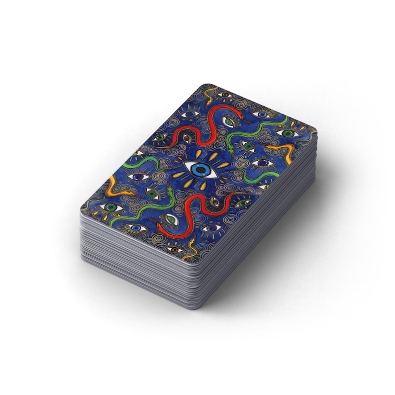 May include: A deck of tarot cards with a blue background and a repeating pattern of eyes and snakes. The center of the deck features a large eye with rays of light. The edges of the cards are silver.