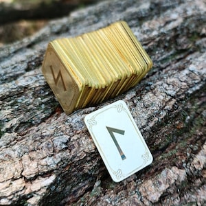 Może przedstawiać: Stos mosiężnych płytek runicznych z jedną płytką pokazującą runę "Fehu" na drewnianej powierzchni.