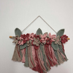 May include: A handmade macrame wall hanging featuring crocheted flowers and leaves in shades of pink, red, and green. The floral arrangement is attached to a wooden dowel and hangs from a metal triangle.