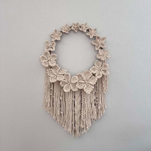 May include: A macrame wall hanging with a circular base and a fringe of tassels. The base is decorated with crocheted flowers in a cream colour.