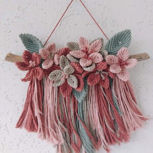 May include: A macrame wall hanging featuring a floral design. The piece showcases pink, dusty rose, and gray flowers and leaves, mounted on a natural wood branch. The bottom has long, fringed strands in coordinating colors.