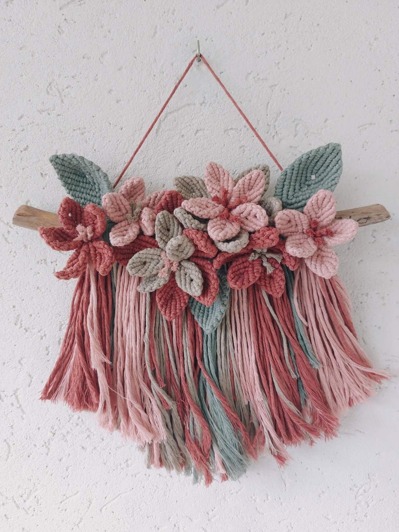 May include: A macrame wall hanging featuring a floral design. The piece showcases pink, dusty rose, and gray flowers and leaves, mounted on a natural wood branch. The bottom has long, fringed strands in coordinating colors.