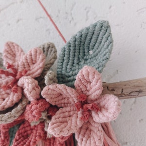 May include: A handmade macrame wall hanging featuring pink and gray flowers and a green leaf. The flowers are attached to a piece of driftwood. The wall hanging is suspended by a pink string.