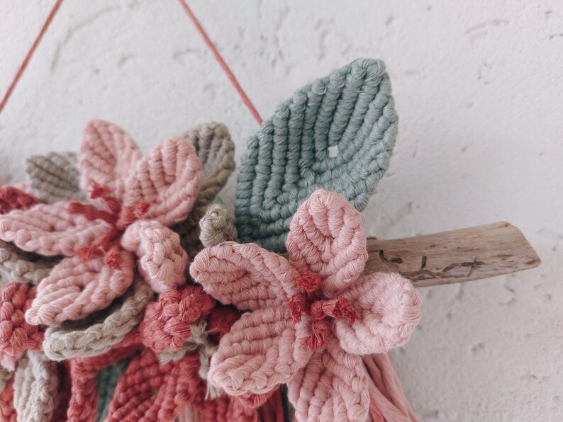 May include: A handmade macrame wall hanging featuring pink and gray flowers and a green leaf. The flowers are attached to a piece of driftwood. The wall hanging is suspended by a pink string.