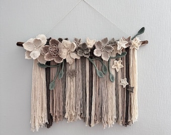 May include: A handcrafted wall hanging featuring a wooden branch adorned with crocheted flowers in shades of cream, brown, and gray. Below, long strands of yarn in similar colors create a textured, fringed effect. The piece is suspended by a thin, white string.