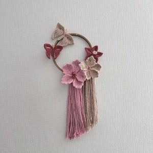 以下が含まれることがあります： ピンクとベージュの糸でできた花と蝶がついた木製フープ。花には長い房が下からぶら下がっています。