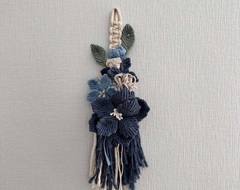 May include: A blue and white macrame wall hanging with a tassel. The wall hanging features a floral design with leaves and flowers.