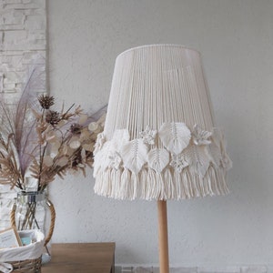 Puede incluir: Una pantalla de lámpara de macramé blanca con ribete de flecos y diseños de hojas. La pantalla de la lámpara está unida a un poste de madera.