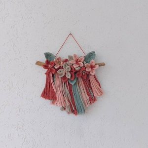 May include: A handmade macrame wall hanging featuring a wooden branch, adorned with fabric flowers in shades of red, pink, and beige, and green leaves. The bottom has a fringe of red, pink, and teal yarn. The wall hanging is suspended by a red string.