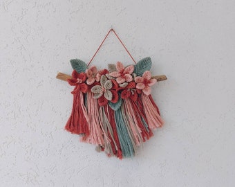May include: A handmade macrame wall hanging featuring a wooden branch, adorned with fabric flowers in shades of red, pink, and beige, and green leaves. The bottom has a fringe of red, pink, and teal yarn. The wall hanging is suspended by a red string.