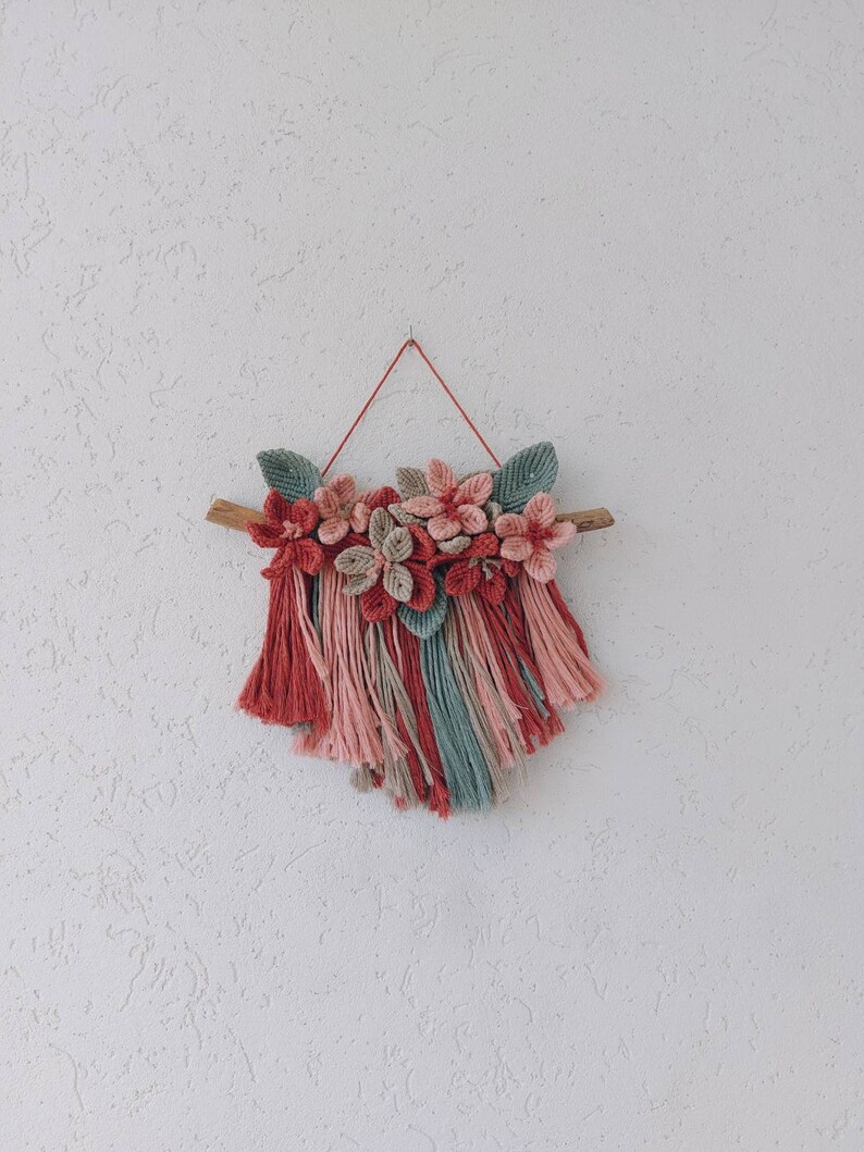 May include: A handmade macrame wall hanging featuring a wooden branch, adorned with fabric flowers in shades of red, pink, and beige, and green leaves. The bottom has a fringe of red, pink, and teal yarn. The wall hanging is suspended by a red string.