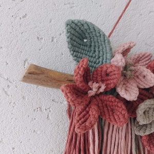 May include: A handmade macrame wall hanging featuring a wooden branch, green leaf, and several flowers in shades of pink and red. The wall decor is mounted on a white textured wall, showcasing the intricate knotting and floral design.