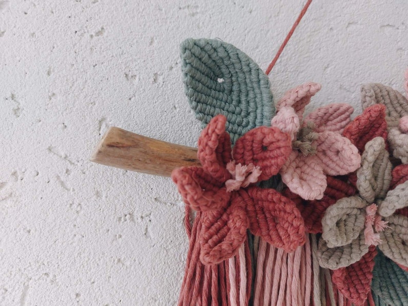May include: A handmade macrame wall hanging featuring a wooden branch, green leaf, and several flowers in shades of pink and red. The wall decor is mounted on a white textured wall, showcasing the intricate knotting and floral design.