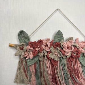 May include: A handmade macrame wall hanging featuring a floral design. The piece incorporates various shades of pink, green, and gray yarn, with a wooden dowel and a metal frame. The textured flowers and leaves add a delicate, artistic touch to the decor.