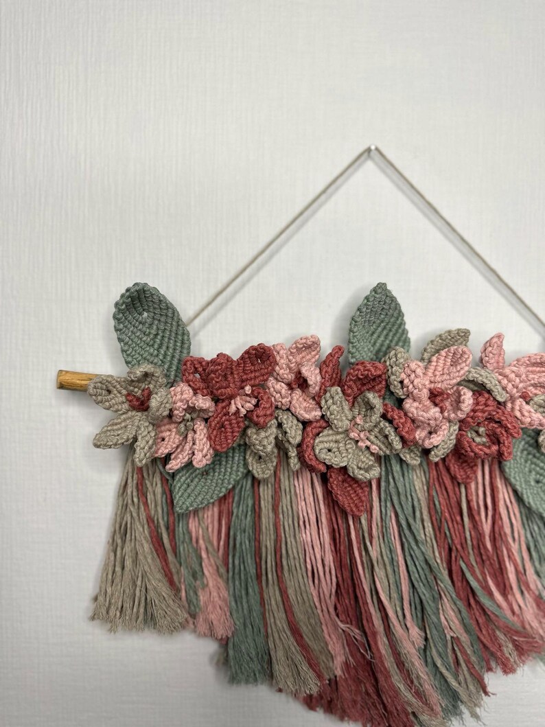 May include: A handmade macrame wall hanging featuring a floral design. The piece incorporates various shades of pink, green, and gray yarn, with a wooden dowel and a metal frame. The textured flowers and leaves add a delicate, artistic touch to the decor.