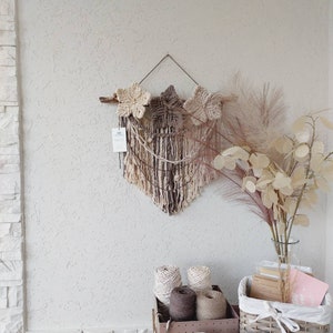 May include: A macrame wall hanging with a brown and beige colour scheme featuring three star shapes. The wall hanging is displayed on a white textured wall with a wicker basket, yarn spools, and candles that spell out "HOME" in front of it.