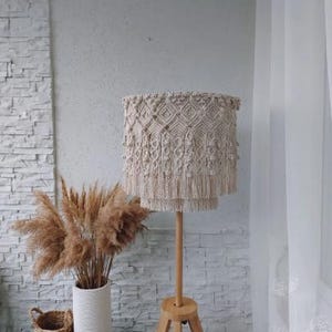 Może przedstawiać: Beżowy abażur z makramy z frędzlami stoi na drewnianej podstawie lampy trójnożnej. Lampa stoi obok białego wazonu z suszoną trawą pampasową. W tle biała ceglana ściana i przezroczysta biała zasłona.