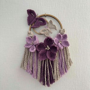 May include: A macramé wall hanging with a wooden ring, three butterfly accents, and three large purple flowers. Below the ring is a fringe of purple, lavender, and beige yarn. This decorative piece is designed for wall display.
