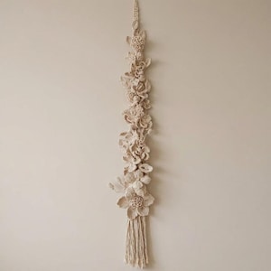 Pode incluir: Uma peça de parede vertical em macramé bege com uma série de flores e folhagens de crochê. O design inclui várias formas florais e uma borla na parte inferior. A peça é montada em uma parede de cor neutra, mostrando um estilo boêmio artesanal.