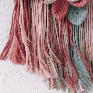 May include: A close-up of a macrame wall hanging with a floral design. The piece features a combination of pink, dusty rose, and gray-green yarn. The textured leaves and fringe add depth to the handmade textile art.