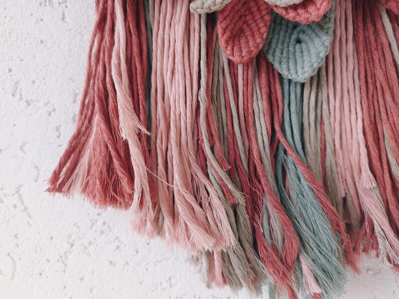 May include: A close-up of a macrame wall hanging with a floral design. The piece features a combination of pink, dusty rose, and gray-green yarn. The textured leaves and fringe add depth to the handmade textile art.