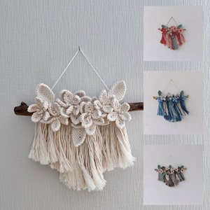 May include: A macrame wall hanging with a cream colored floral design and fringe. The floral design is made of knotted string and the fringe is made of long strands of string.