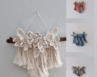 May include: A macrame wall hanging with a cream colored floral design and fringe. The floral design is made of knotted string and the fringe is made of long strands of string.