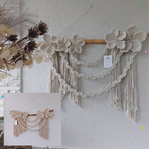 May include: A beige macrame wall hanging with three rows of knotted fringe and two floral designs at the top. The wall hanging is attached to a wooden dowel.