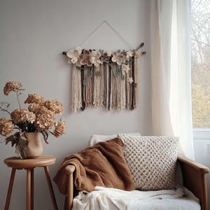 May include: A beige and brown floral wall hanging with a wooden rod and hanging fringe. The wall decor features various sizes of fabric flowers and green leaves. The piece is displayed above a chair with a brown blanket and a cream-colored pillow.
