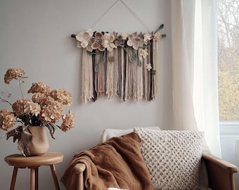 May include: A beige and brown floral wall hanging with a wooden rod and hanging fringe. The wall decor features various sizes of fabric flowers and green leaves. The piece is displayed above a chair with a brown blanket and a cream-colored pillow.