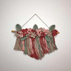 May include: A handmade macrame wall hanging featuring a wooden dowel, a triangle hanger, and a cascade of yarn in shades of red, pink, green, and tan. The top is adorned with crocheted flowers and leaves, creating a textured, floral design.