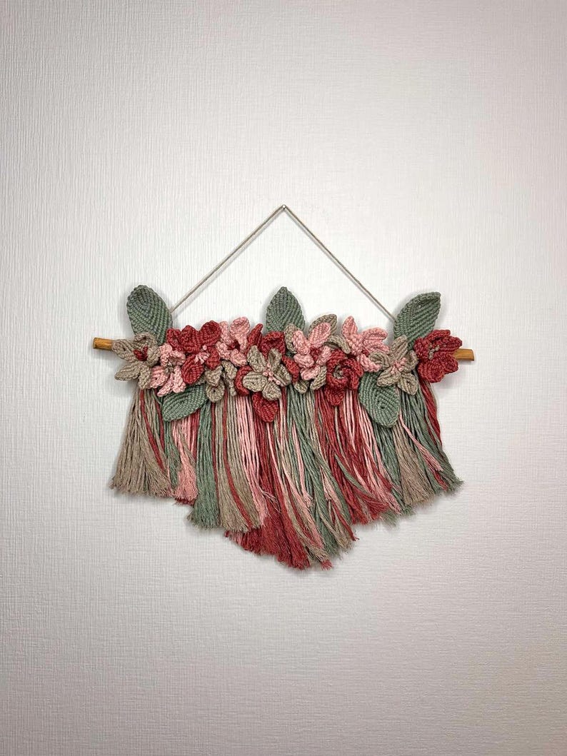 May include: A handmade macrame wall hanging featuring a wooden dowel, a triangle hanger, and a cascade of yarn in shades of red, pink, green, and tan. The top is adorned with crocheted flowers and leaves, creating a textured, floral design.