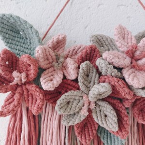 May include: A handmade macrame wall hanging featuring a floral design. The piece showcases flowers in shades of pink and gray, with green leaves and hanging fringe. The wall decor is mounted on a wooden branch and a red string.