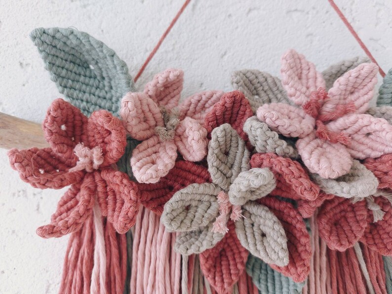 May include: A handmade macrame wall hanging featuring a floral design. The piece showcases flowers in shades of pink and gray, with green leaves and hanging fringe. The wall decor is mounted on a wooden branch and a red string.