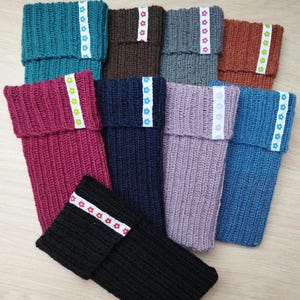 Könnte beinhalten: Sortiment an gestrickten Handyhüllen in verschiedenen Farben mit dekorativem Bandbesatz. Die Hüllen sind aus einem weichen, strukturierten Garn gefertigt und in verschiedenen Farben erhältlich, darunter Teal, Braun, Grau, Orange, Pink, Marineblau, Lila und Schwarz.