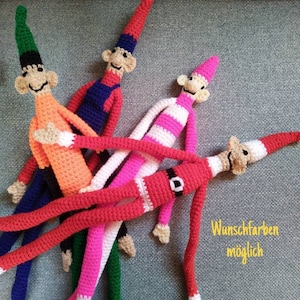 Könnte beinhalten: Vier gehäkelte Elfenpuppen mit langen Gliedmaßen, jede in einer anderen Farbkombination. Die Puppen sind auf einer grauen, strukturierten Oberfläche zu einem Stapel angeordnet. Der Text "Wunschfarben möglich" ist in der unteren rechten Ecke des Bildes sichtbar.