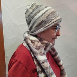 Könnte beinhalten: Ein gestricktes Mützen- und Schal-Set. Die Mütze ist im Beanie-Stil mit horizontalen Streifen in Grau-, Weiß- und Brauntönen gehalten. Der Schal hat ein ähnliches Streifenmuster. Die Person auf dem Bild trägt ein rotes Hemd.