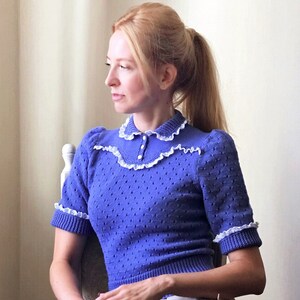 Könnte beinhalten: Ein blauer Strickpullover mit einem weißen Spitzenkragen und -bündchen. Der Pullover hat einen Knopfverschluss am Hals.