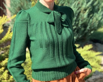 May include: A green knitted sweater with a bow at the neck. The sweater has a ribbed texture and long sleeves.