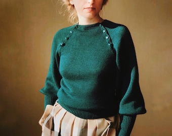 May include: A woman wearing a dark green sweater with buttons on the shoulders and a patterned skirt. She has blonde hair and is wearing a headband.