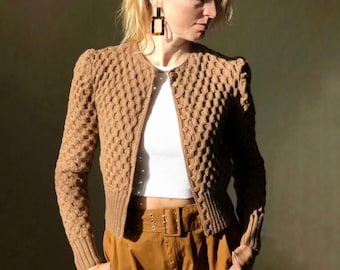 May include: A brown knitted cardigan with a waffle texture, featuring a button-up front and long sleeves. The cardigan is cropped and has a slightly puffed shoulder design. The model is wearing the cardigan with a white t-shirt and brown pants.