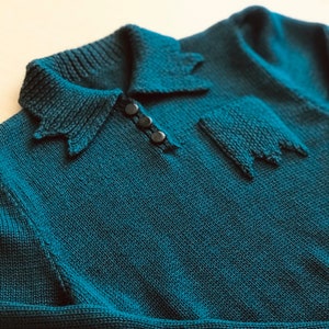May include: A teal blue knitted jumper with a collared neckline and three black buttons. The jumper has a unique design on the front with a knitted pattern.
