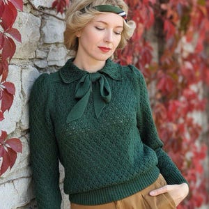 Könnte beinhalten: Ein dunkelgrüner Strickpullover mit einem Bubikragen und Schleifendetail. Der Pullover wird mit einer braunen Hose kombiniert. Ein grün-weißes Stirnband wird getragen. Der Hintergrund zeigt eine Steinmauer und rote Blätter.