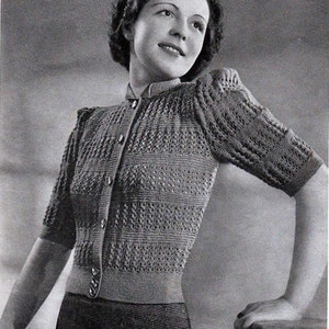 May include: A black and white photo of a woman wearing a knitted cardigan with a button-up front. The cardigan has a delicate openwork pattern and short sleeves. The woman is standing with her right hand resting on a surface.