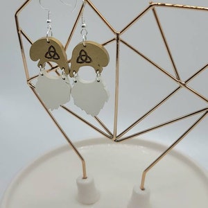 May include: A pair of wooden earrings with a white beard design. The earrings feature a Celtic knot design on the top and are hanging from silver hooks.