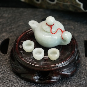 Könnte beinhalten: Ein Miniatur-Tee-Set aus Jade mit einer Teekanne und vier Tassen. Die Teekanne hat eine rote Schnur um den Deckel gebunden. Das Set wird auf einem dunklen Holzständer präsentiert.