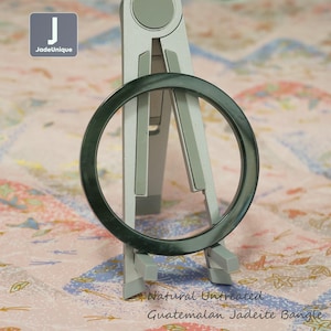 May include: A dark green, circular jadeite bangle. The bangle is displayed on a silver stand against a patterned background. The text "Natural Untreated Guatemalan Jadeite Bangle" is visible.